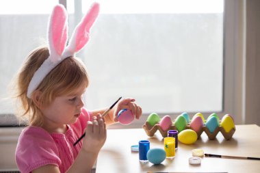 Paskalya tavşanı kulaklı küçük kız renkli yumurta boyuyor. Evde Paskalya Bayramı kutlamaları ve el sanatları konsepti.