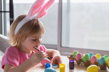 Paskalya tavşanı kulaklı küçük kız renkli yumurta boyuyor. Evde Paskalya Bayramı kutlamaları ve el sanatları konsepti.