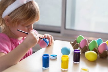 Paskalya tavşanı kulaklı küçük kız renkli yumurta boyuyor. Evde Paskalya Bayramı kutlamaları ve el sanatları konsepti.