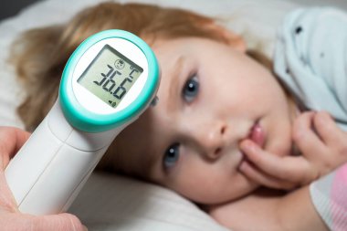 Küçük bir çocuğun ateşini ölçmek, yatakta yatmak. Bir ebeveyn, Coronavirüs salgını sırasında çocuğunun sıcaklığını temassız termometreyle ölçer..