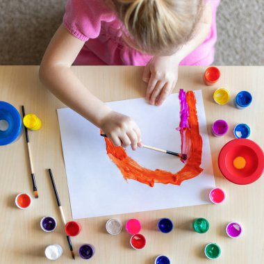 Evdeki masada oturan renkli resimlerle kağıda gökkuşağı resmi çizen küçük kız.