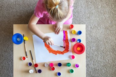 Evdeki masada oturan renkli resimlerle kağıda gökkuşağı resmi çizen küçük kız.