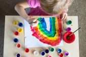 Dívka malování duha na papíře s barevnými barvami sedí u stolu doma