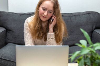 Çalışan genç bir kadın, dizüstü bilgisayarla koltukta oturuyor, telefonla konuşuyor. İş, ev kavramından çalışmak