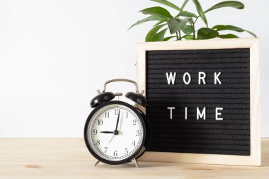 Klasik klasik siyah alarm saati, 'çalışma zamanı' tabelası ve beyaz duvar arkaplan ve metin için alan kopyalayan ahşap masa üzerindeki saksı bitkisi. Zaman kavramı. Yeni çalışma gününün başlangıcı.