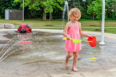 Küçük çocuk sıcak yaz günü yerel halk parkında su ve oyuncaklarla oynuyor. Pembe elbiseli küçük güzel kız çeşme parkında eğleniyor..