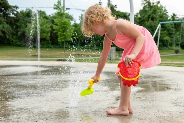 Küçük çocuk sıcak yaz günü yerel halk parkında su ve oyuncaklarla oynuyor. Pembe elbiseli küçük güzel kız çeşme parkında eğleniyor..