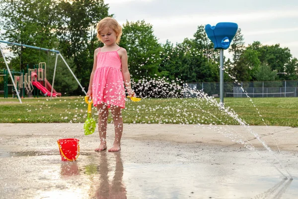 Küçük çocuk sıcak yaz günü yerel halk parkında su ve oyuncaklarla oynuyor. Pembe elbiseli küçük güzel kız çeşme parkında eğleniyor..