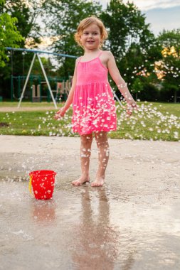 Sıcak yaz günü, yerel halk parkında su ve oyuncaklarla oynayan gülümseyen küçük bir çocuk. Pembe elbiseli küçük güzel kız çeşme parkında eğleniyor..