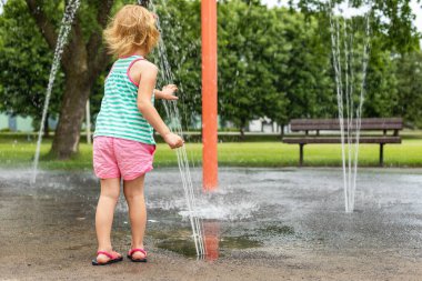 Sıcak bir yaz günü, yerel halk parkında su sıçratma yerindeki suyla oynayan küçük bir çocuk. Küçük güzel kız çeşme bahçesinde eğleniyor..