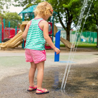 Sıcak bir yaz günü, yerel halk parkında su sıçratma yerindeki suyla oynayan küçük bir çocuk. Küçük güzel kız çeşme bahçesinde eğleniyor..