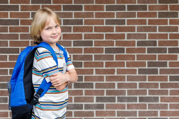 コピースペースのあるレンガの壁に対して学校の建物の近くの美しい笑顔の子供 青いバックパックを持っている小さな金髪の学生 学校のコンセプトに戻る — ストック写真