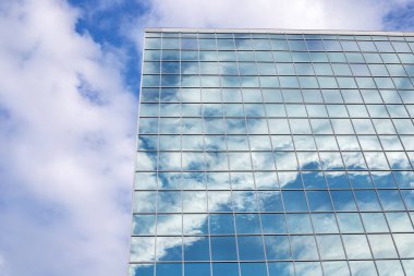 Mavi gökyüzünün yansıması, şehir merkezindeki uzun ve modern cam binada bulutlar.