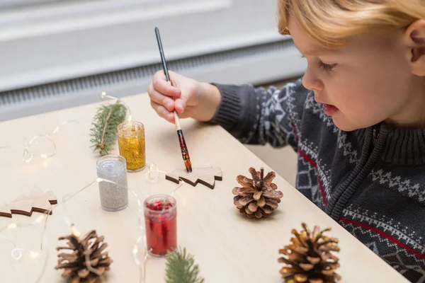 Christmas kids craft activities at home. Little child painting pine cones and wooden Christmas tree decorations. Holiday DIY concept.