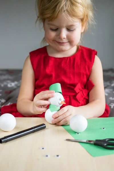 Christmas children craft activities at home. Little smiling girl in red dress making snowman. Kid is busy playing. DIY concept.