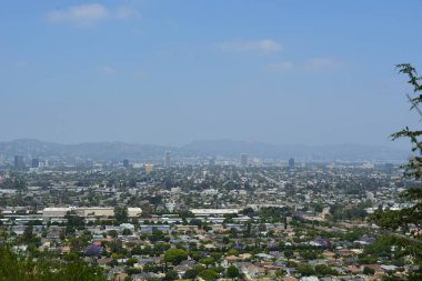 Parlak güneş, evler ve ağaçlarla dolu geniş bir şehrin üzerine ılık bir ışık saçıyor. Dağlar uzaklardan yükselir, berrak mavi bir gökyüzüne karışarak, huzurlu ama canlı bir manzara yaratır..
