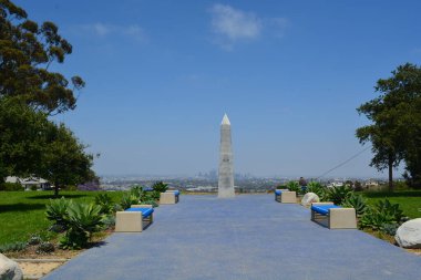 Açık mavi bir gökyüzünün altında, huzurlu bir parkın üzerinde uzun bir anıt yükselir. Çevredeki banklar ziyaretçileri çarpıcı şehir manzarası ve bitkilerle dolu verimli manzaraların tadını çıkarmaya davet ediyor..