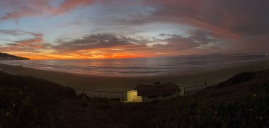 Güneş batarken, gökyüzü renklerle dolup taşar, aşağıdaki kumlu kumsalda sıcak bir parıltı bırakır. Dalgalar yavaşça kıyıya vururken, yakınlardaki evler sönen ışığın tadını çıkararak sakin bir atmosfer yaratıyor..