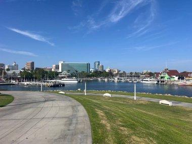 Canlı bir rıhtım sahnesinde, modern binalar ve güneşli bir günde yemyeşil parklarla çevrili marinada sallanan tekneler görülüyor..