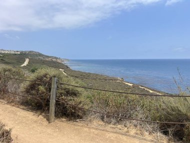 Sahil kenarındaki dolambaçlı bir yol sakin denizi ve yeşil tepeleri nefes kesen bir manzara sağlar. Güneş parıldıyor, sudan yansıyor ve doğa yakınlarda gelişiyor..