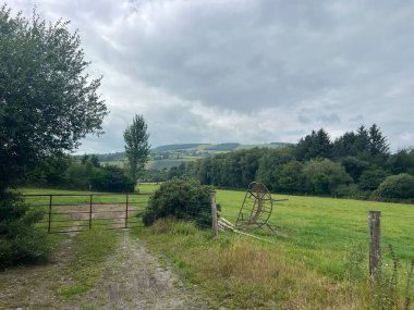 Çerçevelenmiş ağaçlar ve kırsal bir kapı ile uzak kayan tepelere doğru yemyeşil tarlalar uzanıyor. Bulutlu gökyüzü, yaklaşan hava değişikliğine işaret ediyor. Sahneye dingin bir atmosfer katıyor..