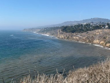 Güneş ışığı suyun üzerinde parıldarken, yumuşak dalgalar altın kuma çarpar. Uzaktan yükselen kayalıklar, yeşilliklerle kaplı, huzurlu bir kıyı manzarası sergiliyor..