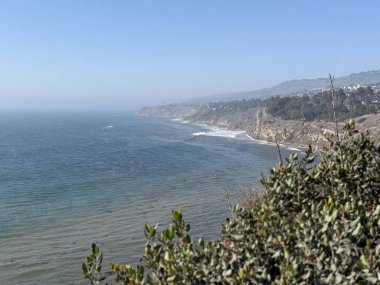 Uçsuz bucaksız okyanus, kumlu kıyılara doğru yumuşak dalgalarla dalgalanan berrak bir gökyüzü altında uzanıyor. Bereketli yeşillik, uçurum boyunca manzarayı çerçeveler ve huzurlu bir manzara yaratır..