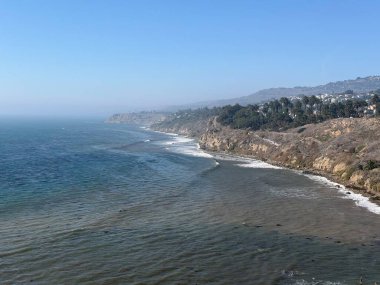 Dalgalar yavaşça kıyıya vururken, uçurumlar kıyı şeridi boyunca dramatik bir şekilde yükseliyor. Yeşil tepeler ve açık mavi gökyüzü bu sakin manzarayı tamamlıyor..