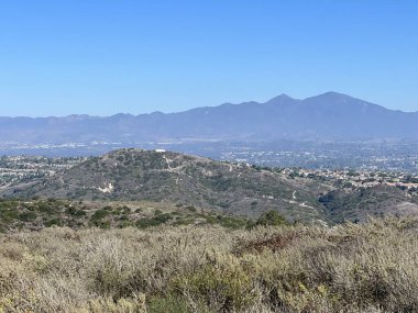 Arka plandaki görkemli dağlara zıt olarak altın bitki örtüsüyle kaplanmış, yuvarlanan tepeleri gösteren nefes kesici bir manzara. Gökyüzü geniş ve berrak uzanıyor, güzelliği arttırıyor..