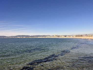 Açık mavi gökyüzü sakin suların üzerine uzanır, kıyı manzarasını yansıtır. Uzakta kumlu sahil uzanır, ve yumuşak dalgalar kıyıya vurur, deniz kenarında huzurlu bir geri çekilme yaratır..