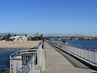 İnsanlar, parlak mavi gökyüzünün altında, köpüklü su ve çarpıcı dağlarla çevrili iskelede sakin bir yürüyüşün tadını çıkarıyorlar. Sahil ve kasaba kıyısı. Resimli bir sahne sunuyor..
