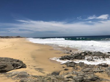 Sakin bir sahil sahnesi, altın kumlara yuvarlanan hafif dalgaları yakalar. Güneş, kıyı boyunca kayalık oluşumları aydınlatan parlak bir şekilde tepemizde parlıyor. Okyanus kıyısında huzurlu bir tatil..