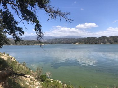 Berrak bir manzara, yemyeşil ve uzak tepelerle çevrili gölün parıldayan suyunu yakalar. Gökyüzü aydınlık ve berrak, öğleden sonra huzur ve doğal güzellik sunuyor..