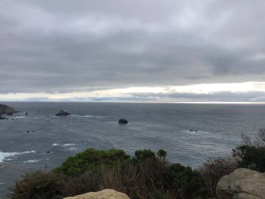 Güneş kalın bulutların ardında batarken dalgalar yavaşça kayalık çıkıntılara çarpar. Sakin deniz bastırılmış ışığı yansıtır ve kıyı şeridinde sakin bir atmosfer yaratır..