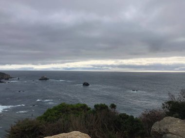 Güneş batarken dalgalar engebeli kayalara hafifçe çarparak sakin okyanusun üzerine yumuşak bir ışık saçar. Ufukta kara bulutlar toplanıyor ve sakin bir atmosfer yaratıyor..