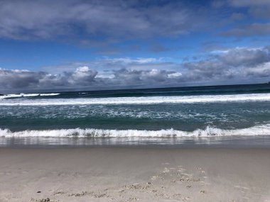 Yumuşak dalgalar el değmemiş kumlu bir plaja yuvarlanarak huzurlu bir ortam yaratıyor. Canlı mavi gökyüzü ve dağınık bulutlar bu kıyı cennetinin huzurlu atmosferini güçlendirir..