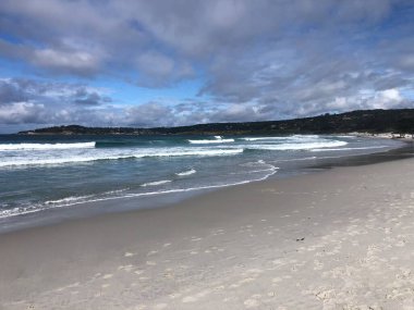 Sakin bir kıyı şeridi sıcak kuma vuran yumuşak dalgaları yansıtır. Yukarıdaki bulutlu gökyüzü rahatlamak ve kaçmak için ideal sakin bir atmosfer yaratıyor..