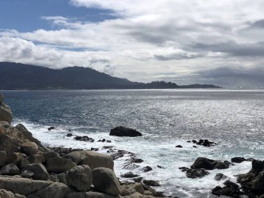 Dalgalar sakin bir sahil şeridi boyunca engebeli kayalara hafifçe çarpar. Uzak dağlar bulutlu bir gökyüzünde solup doğal güzelliklerle dolu huzurlu bir sahil manzarası yaratıyor..