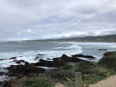 Bulutlu bir günde, dalgalar kıyıdaki karanlık kayalara çarpar ve büyüleyici bir manzara yaratır. Okyanus ufka kadar uzanıyor, izleyicileri güzelliğini keşfetmeye çağırıyor..