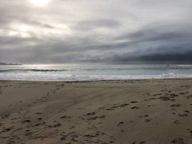 Yumuşak dalgalar, gri bulutların altında sakin bir kıyı şeridine çarpar. Akşam ışığı huzurlu bir atmosfer yaratır, sakin bir yansıma ve rahatlamaya yol açar..