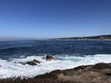 Parlak güneş ışığı, kayalık oluşumlara çarptıklarında okyanus dalgaları üzerinde dans eder. Kıyı şeridi arka planda uzanır, deniz kenarında güzel bir günde huzurlu bir manzara sunar..