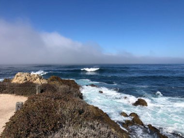 Dalgalar engebeli kayalara çarpıyor, okyanusun üzerinde hafif bir sis gibi. Parlak mavi gökyüzü ve kıyı şeridi yansıma ve kaçma için mükemmel sakin bir atmosfer yaratır..