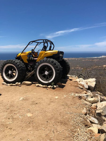 A bright yellow off-road vehicle sits confidently on a rocky ledge with stunning ocean views. The clear blue sky enhances the adventurous atmosphere and natural beauty.