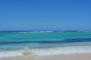 Dalgalar yumuşak kumlu sahilde yavaşça dolaşırken parlak güneş canlı bir turkuaz denizin üzerinde parlıyor. Kumsal huzur verici, doğanın güzelliğinin rahatlatıcı ve keyifli..