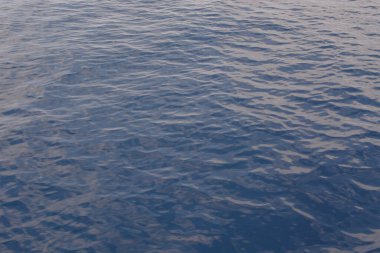 Sakin sular durgun gökyüzünü ve güneş ışığını yansıtıyor. Yumuşak dalgalar rahatlatıcı bir atmosfer yaratır, deniz kenarında huzur ve rahatlamaya davetiye çıkarır..