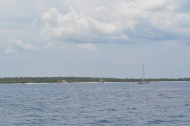 Parlak mavi okyanus suları yemyeşil bir tropikal adaya doğru uzanıyor. Bir yelkenli, etrafını palmiye ağaçları ve yumuşak bulutların sardığı kıyı şeridinde sorunsuzca yol alır..