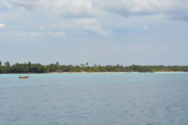 Küçük ahşap bir tekne parçalı bulutlu bir gökyüzünün altında yemyeşil bir kıyı şeridi tarafından çerçevelenmiş sakin sularda usulca hareket eder. Bu huzurlu sahne doğanın güzelliğini yansıtıyor..