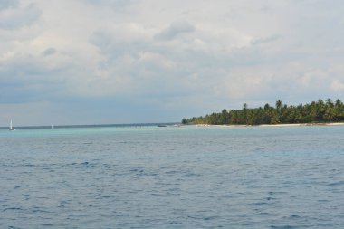 Dalgalanan gök mavisi sular sallanan palmiyelerle süslenmiş uzak bir kıyı şeridine doğru uzanıyor. Huzurlu bir atmosfer sarar bu kıyı cennetini bulutlar usulca yükselirken.