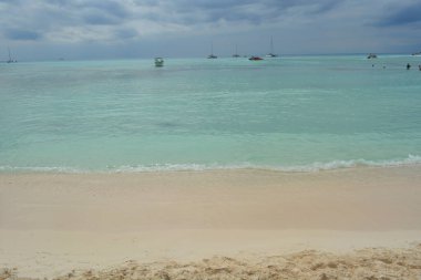 Sakin sular yumuşak kumlu sahilde yavaşça dolaşırken, tekneler turkuaz denizde sorunsuzca süzülüyor. Gökyüzü bulutlu, dinlenmek için mükemmel sakin bir atmosfer yaratıyor..