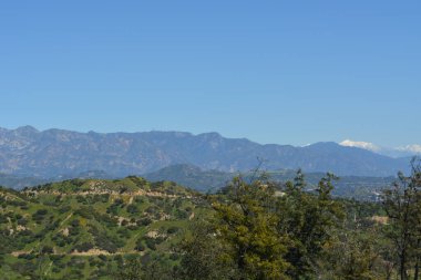 Güneşli bir günde, geniş bir dağ sırasının nefes kesici manzarası. Önünde yemyeşil tepeler ve uzak tepeler karla kaplanmış. Doğanın kucağında huzurlu bir kaçış.